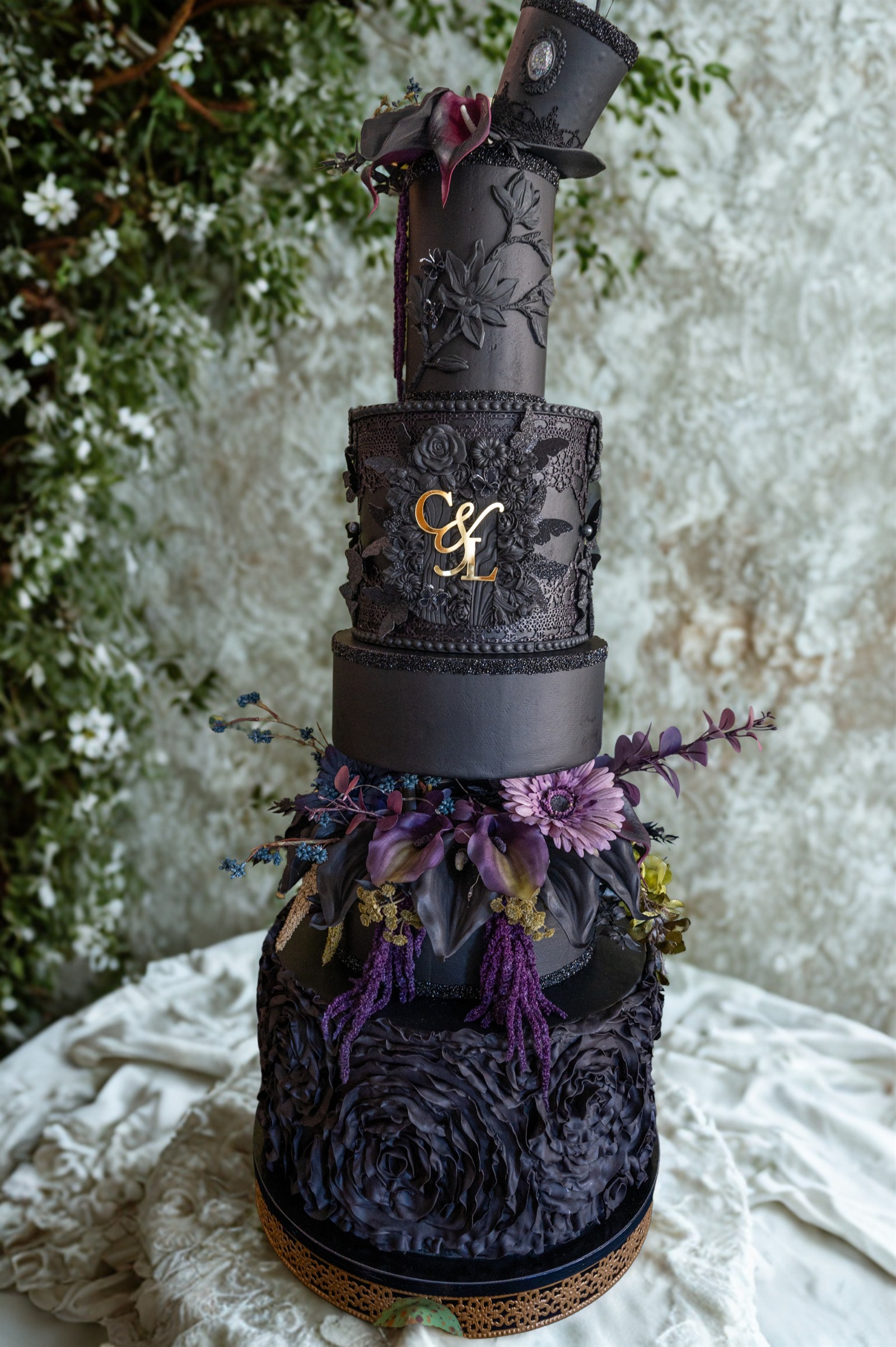 Tall sculptural black celebration cake decorated with flowers, foliage and gold initials